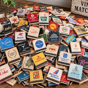 May include: A large collection of rectangular vintage matchbooks featuring various colorful brand logos and text, including names like Ramada Inn, Holiday Inn, Shell, Pan Am, TWA, Caesars Palace, and Have a Coke, piled together on a wooden surface.