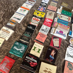May include: A collection of vintage matchbooks with various designs and text, including logos, illustrations, and advertisements. The matchbooks are arranged on a wooden surface, showcasing a variety of colors and styles. Some matchbooks feature text like "Golden Nugget" and "Occidental".