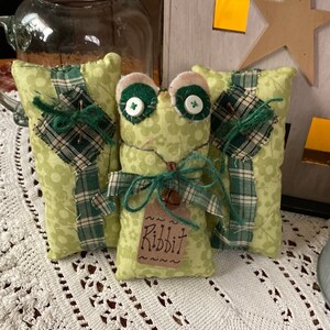 May include: A handmade frog-shaped plush toy with button eyes and a tag that says "Ribbit." The frog is light green with a floral pattern, and it has a green bow and a small bell. Two matching green pillows with plaid accents flank the frog.