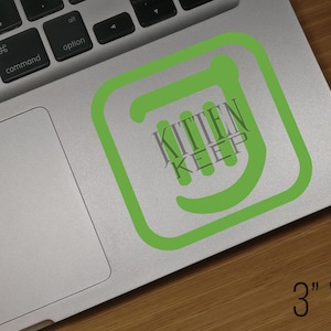 Könnte beinhalten: Ein grüner und weißer Aufkleber mit dem Text "Kitten Keep" auf einem silbernen Laptop. Der Aufkleber ist 7,6 cm breit.