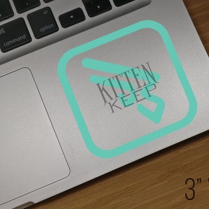 May include: A teal blue sticker with a white outline and the text "Kitten Keep" in a white sans-serif font. The sticker is on a silver laptop.