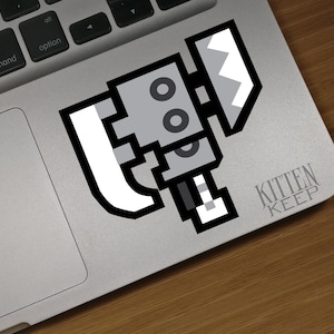 May include: A gray and white pixelated robot holding a white axe with a black handle. The robot has two black eyes and a black mouth. The robot is on a silver laptop with the text "KITTEN KEEP" in black letters.