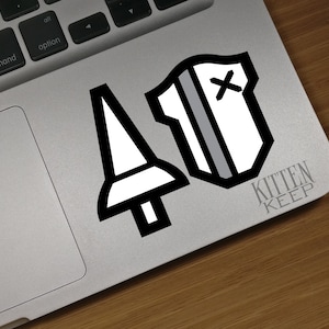 May include: A black and white sticker with a sword and a shield with a cross on it. The sticker is on a silver laptop. The text "Kitten Keep" is below the sticker.