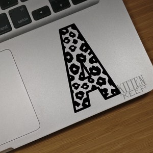 May include: A black and white leopard print letter A sticker on a silver laptop. The sticker says "Kitten Keep" in the bottom right corner.