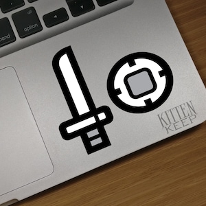 May include: A white sticker with a black outline of a sword and a target with a gray center. The sticker is on a silver laptop with the text "KITTEN KEEP" below the sticker.