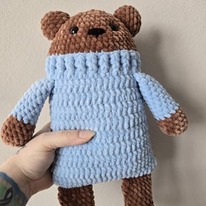 May include: A handmade brown and blue crochet bear. The bear has a brown head, ears, arms, and legs. It is wearing a light blue sweater. The bear has black eyes and a black nose. The bear is soft and plush.