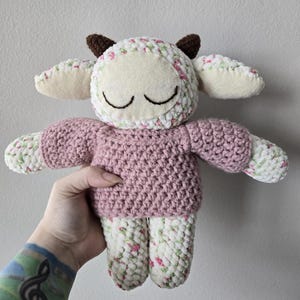May include: A handmade crochet sheep doll with a pink sweater and floral patterned legs and head. The sheep has brown horns and closed eyes, giving it a sleepy appearance. The doll is soft and cuddly.