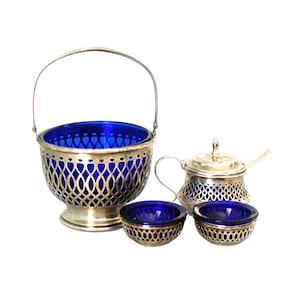 Op de afbeelding: Een zilveren suiker- en roomset met blauwe glazen voeringen. De roomkan heeft een deksel en een kleine lepel. De suikerpot heeft een handvat en een decoratief opengewerkt ontwerp.