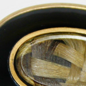 May include: A gold-toned ring with a black enamel band and a clear glass oval inset containing a woven lock of hair.