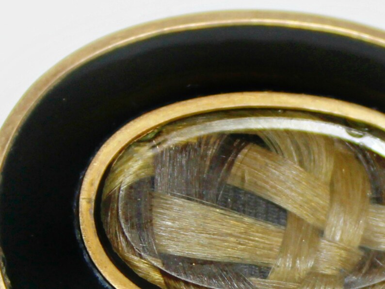 May include: A gold-toned ring with a black enamel band and a clear glass oval inset containing a woven lock of hair.