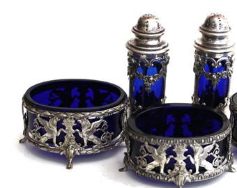 May include: A set of two cobalt blue glass bowls and two matching shakers, each encased in intricate silver-toned metal filigree featuring winged lion motifs and standing on small decorative feet, with perforated silver-toned metal lids.