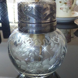 May include: A clear glass bottle with a silver lid and etched floral designs. The bottle has a silver top with an intricate floral design.