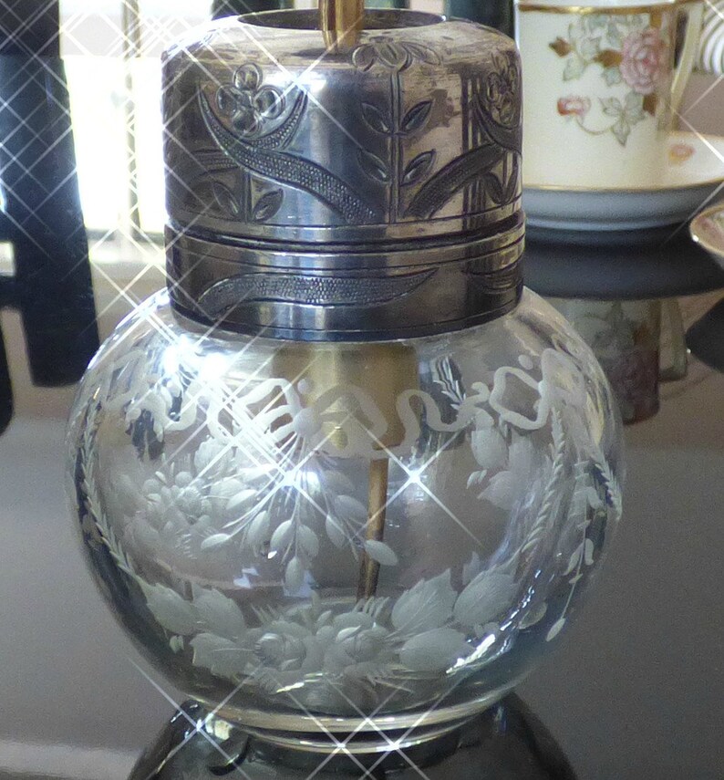May include: A clear glass bottle with a silver lid and etched floral designs. The bottle has a silver top with an intricate floral design.