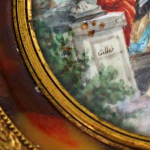 Pu&ograve; includere: Un primo piano di un dipinto in miniatura incorniciato in oro. Il dipinto raffigura una scena con figure in un ambiente da giardino. La firma "Gillet" &egrave; visibile su una struttura in pietra nel dipinto.