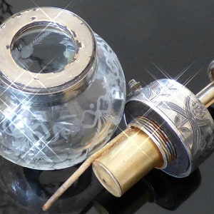 May include: A silver and glass bottle stopper with an etched floral design. The stopper has a clear glass bulb with a silver top and a brass pump.