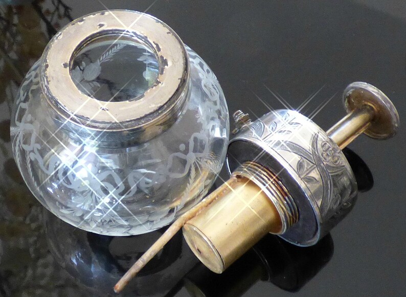 May include: A silver and glass bottle stopper with an etched floral design. The stopper has a clear glass bulb with a silver top and a brass pump.