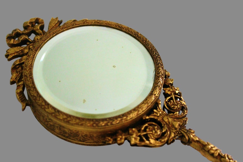 Pu&ograve; includere: Uno specchio da mano dorato con una cornice e un manico decorati. Lo specchio ha una forma circolare e una superficie in vetro trasparente.