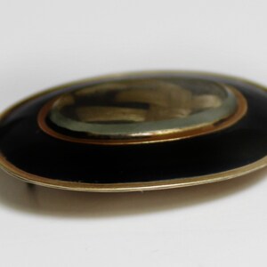 May include: An oval-shaped brooch with a black enamel background and a gold-toned rim. The center of the brooch features a glass dome with a gold-toned design.