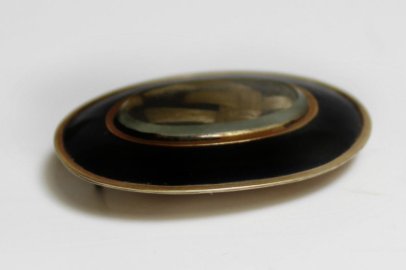 May include: An oval-shaped brooch with a black enamel background and a gold-toned rim. The center of the brooch features a glass dome with a gold-toned design.
