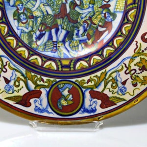May include: A large, round ceramic plate with a colorful design. The center of the plate features a scene of people dancing, while the outer rim is decorated with intricate floral patterns in shades of blue, yellow, and red.