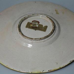 May include: The underside of a white ceramic plate with a yellow rim. A small, rectangular, paper label is attached to the center of the plate.