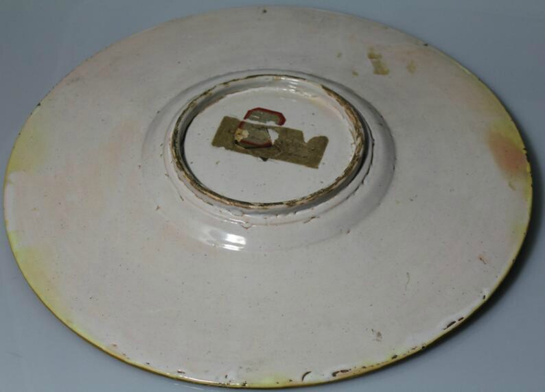 May include: The underside of a white ceramic plate with a yellow rim. A small, rectangular, paper label is attached to the center of the plate.
