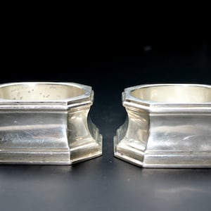 May include: Two silver-colored napkin rings. Each ring has an octagonal top and a flared base. The rings are made of a shiny metal and sit on a dark surface. The napkin rings are designed to hold cloth napkins.