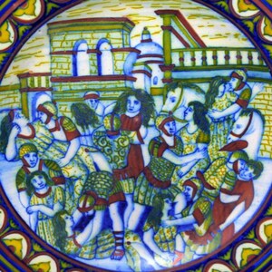 May include: A ceramic plate with a colorful scene of people in a medieval setting. The plate has a blue and white border with yellow and red accents.