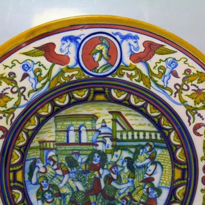 May include: A ceramic plate with a colorful design. The center of the plate features a scene of people in a town square. The border of the plate is decorated with floral motifs and winged horses.