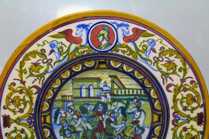 May include: A ceramic plate with a colorful design. The center of the plate features a scene of people in a town square. The border of the plate is decorated with floral motifs and winged horses.