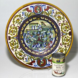 May include: A large, round, ceramic plate with a colorful, intricate design. The plate features a central scene of people in a city setting, surrounded by floral motifs. The plate has a yellow, red, blue, and green color scheme.