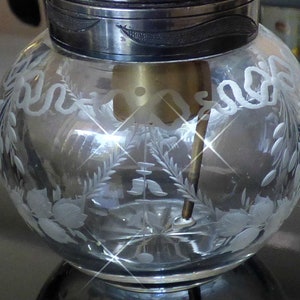 May include: A clear glass jar with a silver lid and etched floral designs. The jar has a small opening at the top and a metal rod extending from the bottom.