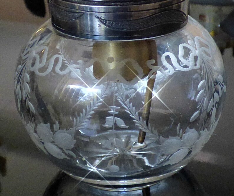May include: A clear glass jar with a silver lid and etched floral designs. The jar has a small opening at the top and a metal rod extending from the bottom.