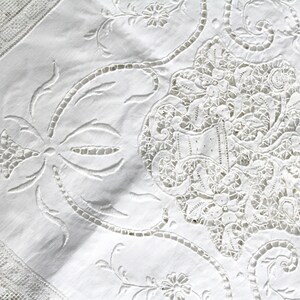 May include: White cotton tablecloth with intricate embroidered lace detailing. The design features a central floral motif with delicate leaves and swirls.