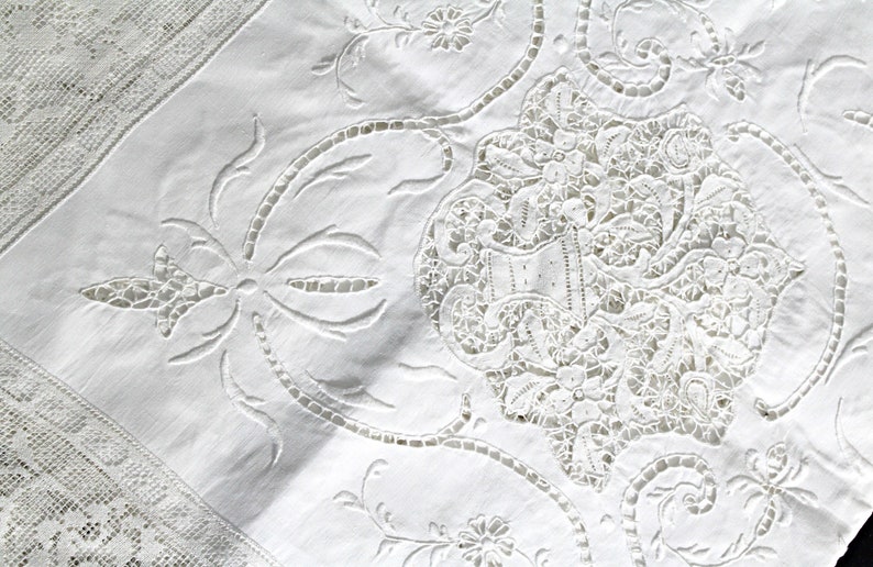 May include: White cotton tablecloth with intricate embroidered lace detailing. The design features a central floral motif with delicate leaves and swirls.