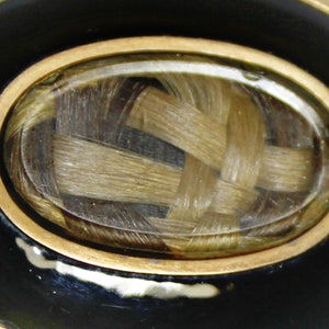 May include: A gold-rimmed oval locket with a black enamel background. Inside the locket is a woven braid of light brown hair.