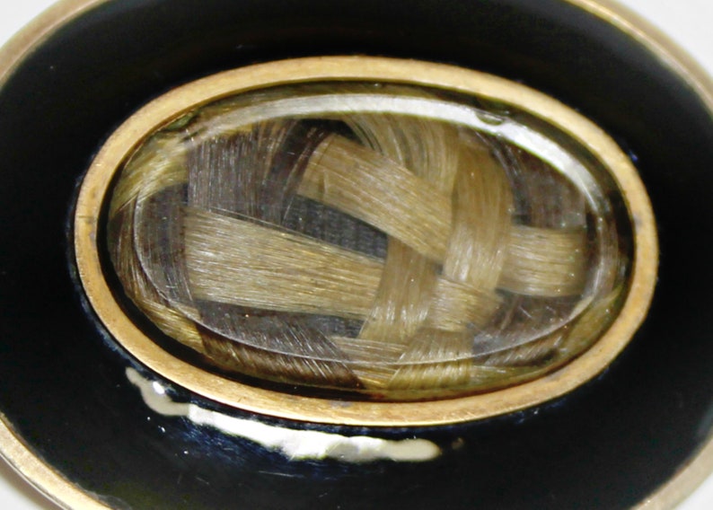 May include: A gold-rimmed oval locket with a black enamel background. Inside the locket is a woven braid of light brown hair.