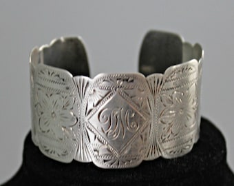 Puede incluir: Brazalete de plata con un diseño floral y geométrico grabado. El panel central presenta una forma de diamante con iniciales grabadas. El brazalete tiene un borde festoneado y se muestra sobre una superficie negra.