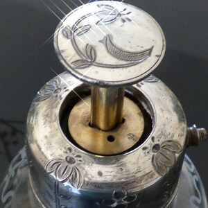 May include: Close-up of a silver bottle stopper with a decorative floral design. The stopper has a brass rod and a circular top with a floral design.