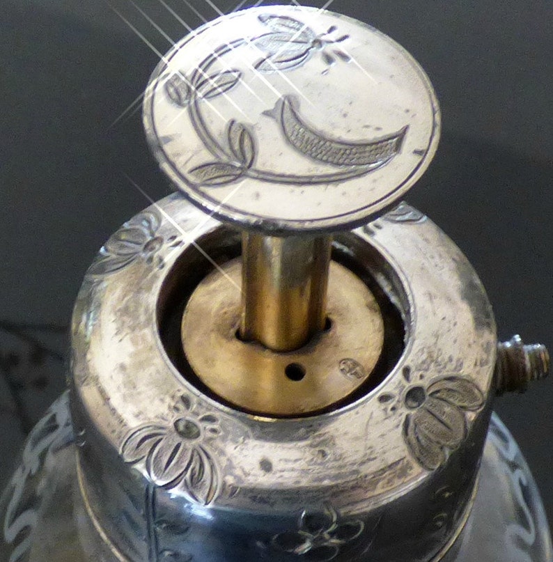May include: Close-up of a silver bottle stopper with a decorative floral design. The stopper has a brass rod and a circular top with a floral design.