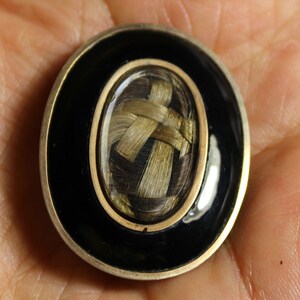 May include: A small oval locket with a black enamel border and a gold trim. Inside the locket is a woven braid of hair.