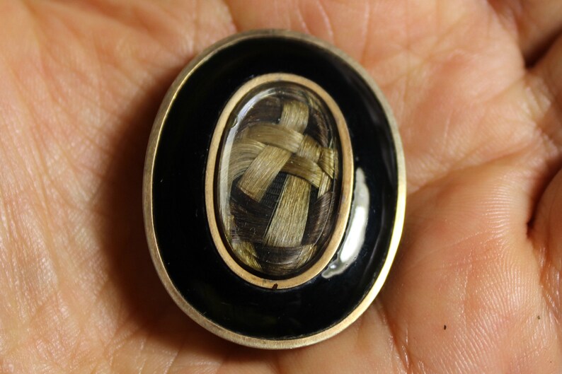 May include: A small oval locket with a black enamel border and a gold trim. Inside the locket is a woven braid of hair.