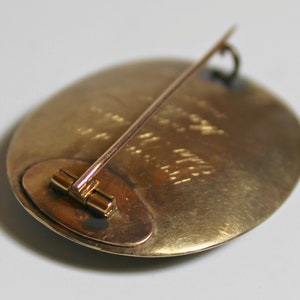 May include: A gold-toned oval brooch with a pin on the back. The brooch has a textured surface and a small, round, metal piece on the back.