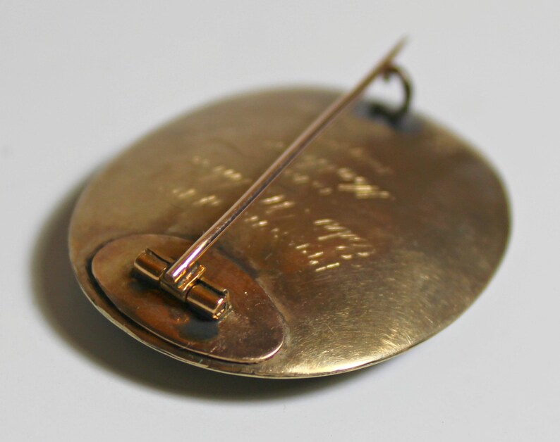 May include: A gold-toned oval brooch with a pin on the back. The brooch has a textured surface and a small, round, metal piece on the back.