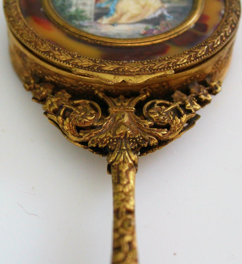 Pu&ograve; includere: Uno specchio da mano dorato con un manico decorato a volute e una superficie rotonda a specchio. Lo specchio ha un bordo decorativo a festoni e una piccola scena dipinta al centro.