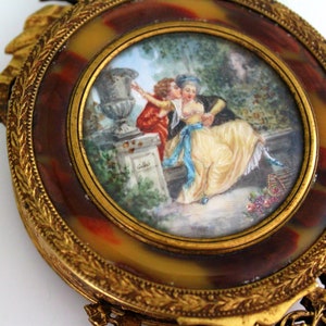 Pu&ograve; includere: Una cornice dorata ornata con un cammeo che rappresenta una scena romantica. Il cammeo mostra una coppia in un giardino con un vaso e una gabbia per uccelli. La cornice &egrave; decorata con intricati motivi floreali.