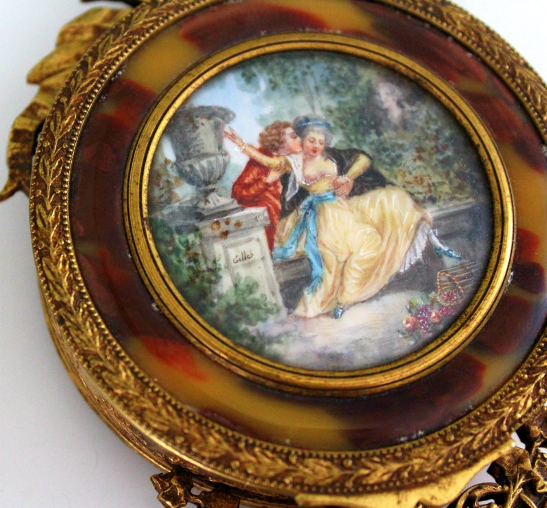 Pu&ograve; includere: Una cornice dorata ornata con un cammeo che rappresenta una scena romantica. Il cammeo mostra una coppia in un giardino con un vaso e una gabbia per uccelli. La cornice &egrave; decorata con intricati motivi floreali.