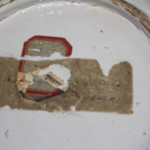May include: Close-up of a white ceramic plate with a faded, partially torn label on the bottom. The label has red, blue, and black lettering and the text "Napier & Son, London" is visible.