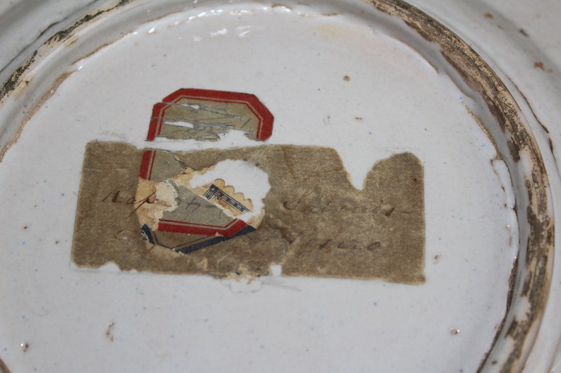 May include: Close-up of a white ceramic plate with a faded, partially torn label on the bottom. The label has red, blue, and black lettering and the text "Napier & Son, London" is visible.