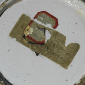 May include: Close-up of a white ceramic plate with a brown paper label that says "14-1-14" and a red and blue label with a faded image.